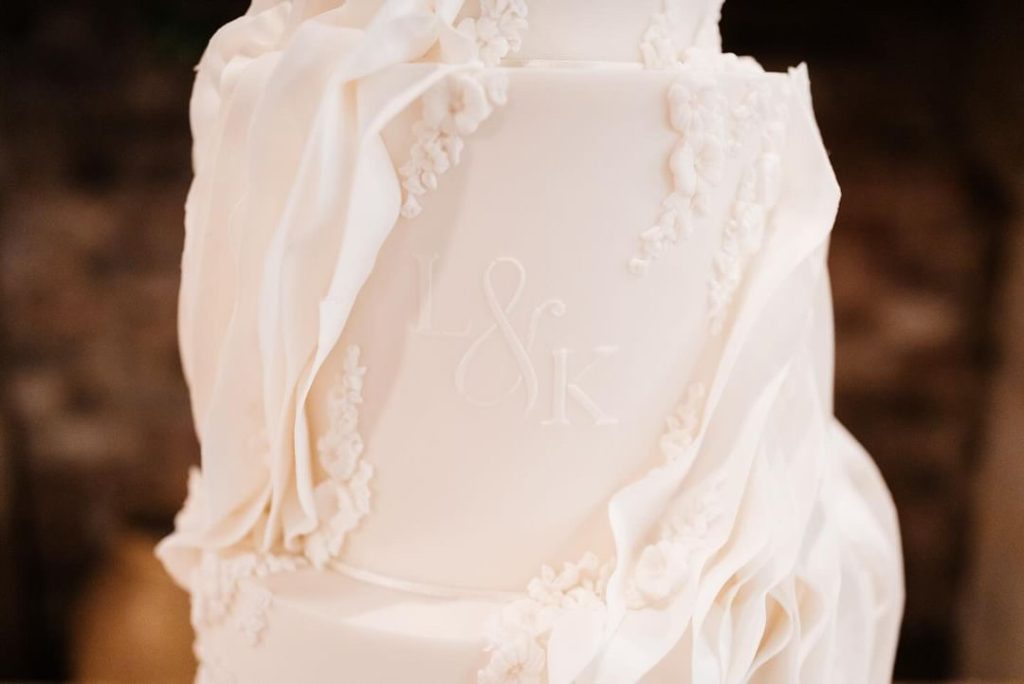 A close-up of a white tiered wedding cake decorated with draped fondant and floral details, with the initials "L & K" embossed in the center.