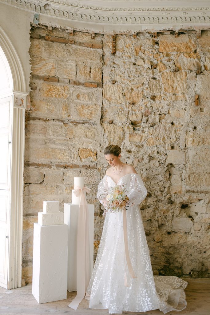 Bride in a white floral gown holding a bouquet, standing beside a set of three-tier white wedding cakes against a rustic stone wall backdrop.
