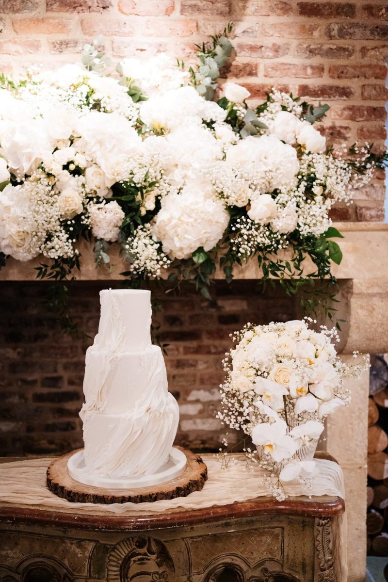 A three-tier white wedding cake with textured icing sits on a wooden slab. Next to the cake is a bouquet of white flowers and baby's breath. A floral arrangement decorates the brick wall background.