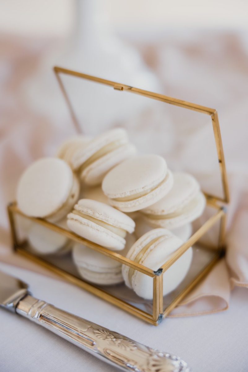 A glass-and-gold box filled with white macarons sits on a table next to an ornate butter knife.