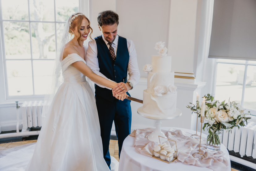A bride and groom in formal attire are cutting a white tiered wedding cake with a knife. A floral arrangement and candles are on the table beside the cake. Created by The Little Cakery, a wedding Cake Designer North East England.