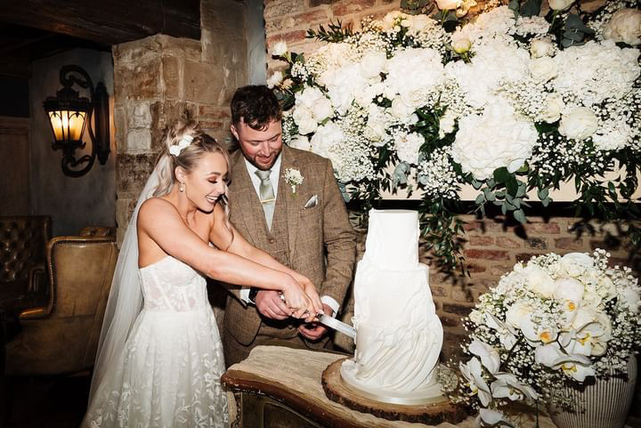 Wedding cake in South Shields. A bride and groom are cutting a white, tiered wedding cake together. They are smiling and surrounded by white floral decorations.