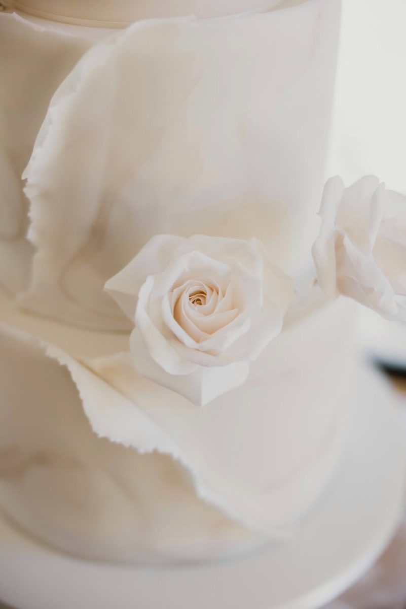 Close-up of a tiered white wedding cake decorated with realistic white rose flowers and delicate, ruffled fondant details.