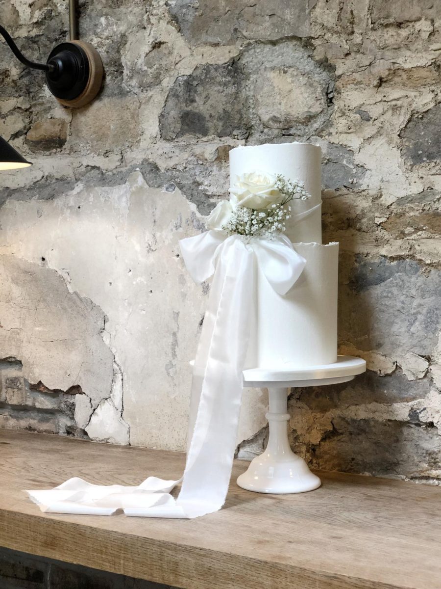 A luxury fondant wedding cake with two-tiers and a white rose and ribbon, placed on a white pedestal stand against a textured stone wall.