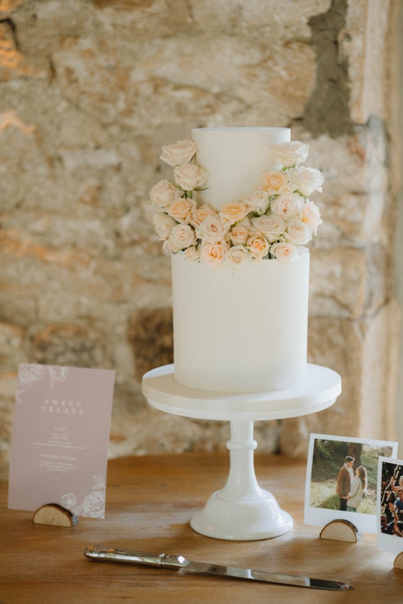 A two-tier white wedding cake with floral decorations on a white stand, accompanied by a wedding invitation and a photo on a wooden table beside a cake knife.