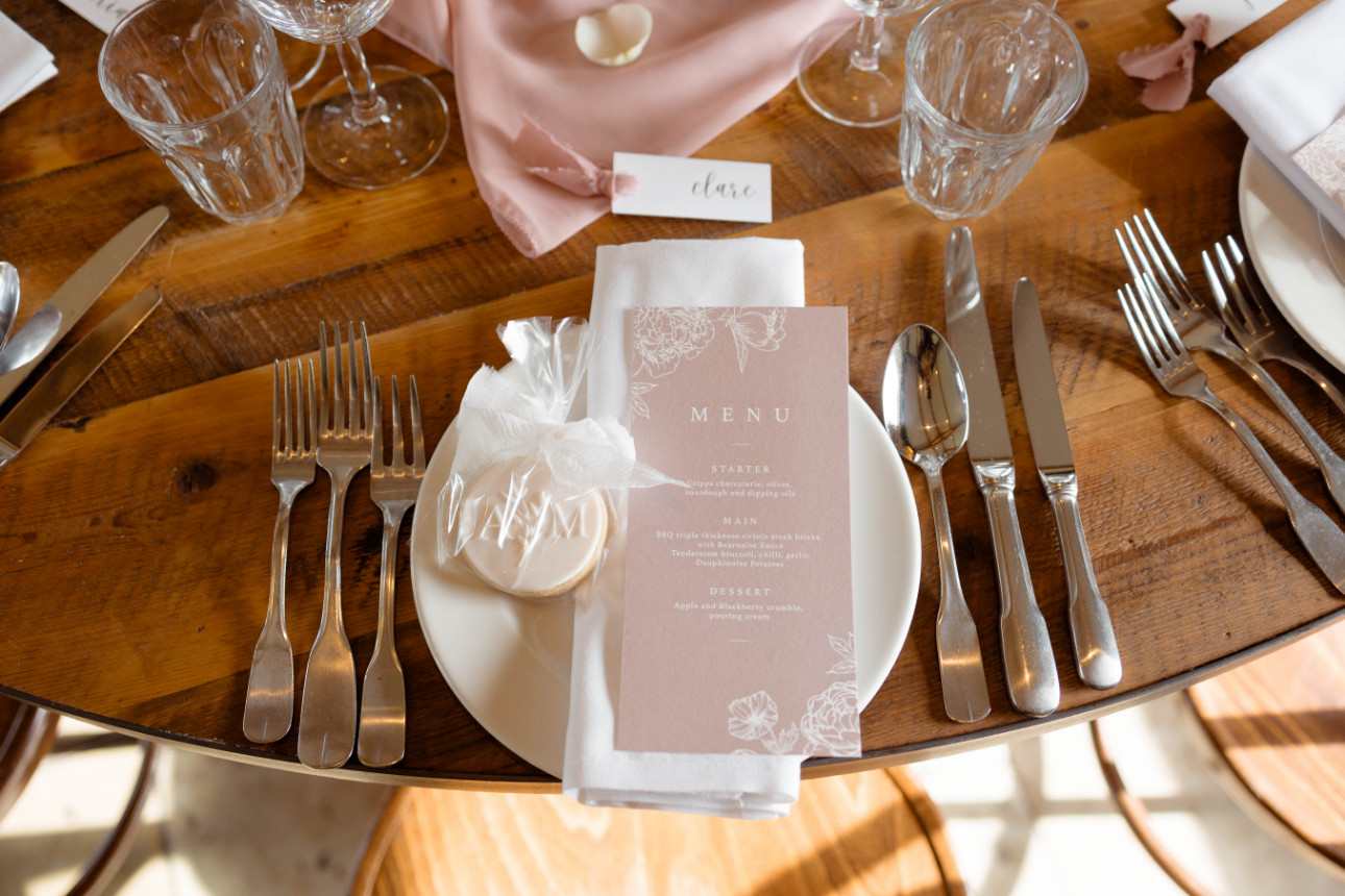 A table setting with a menu, wrapped cookie, napkin, and cutlery on a wooden table. Glassware and a name card are arranged above the plate. Created by The Little Cakery, a wedding Cake Designer North East England.