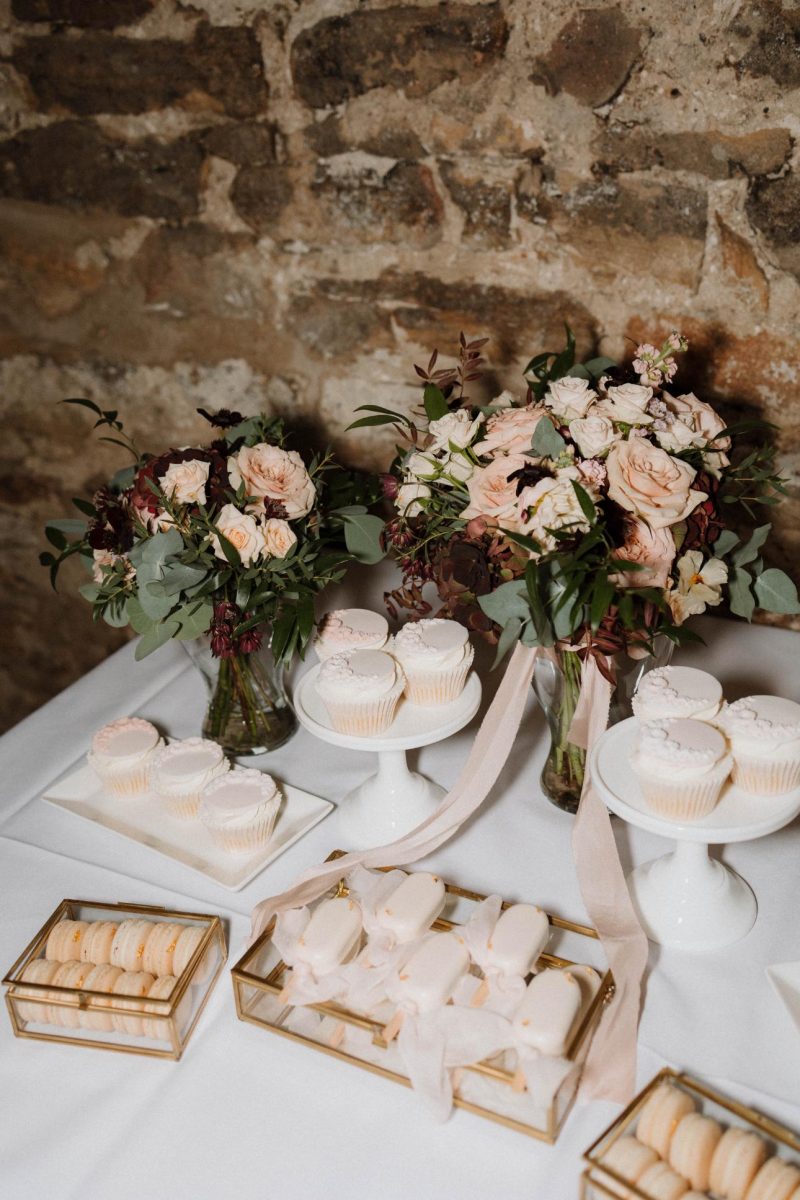 Wedding cake in South Shields. A table decorated with vases of flowers, plates of cupcakes, and trays of macarons against a stone wall backdrop.