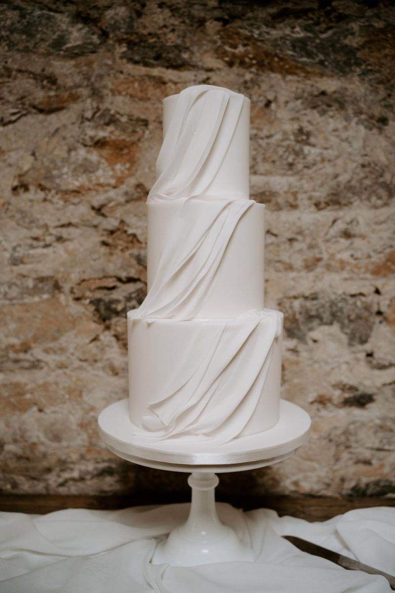 A luxury fondant wedding cake with three-tiers and draped, smooth fondant decorations, displayed on a white stand against a textured stone wall background. Created by The Little Cakery, a wedding Cake Designer North East England.