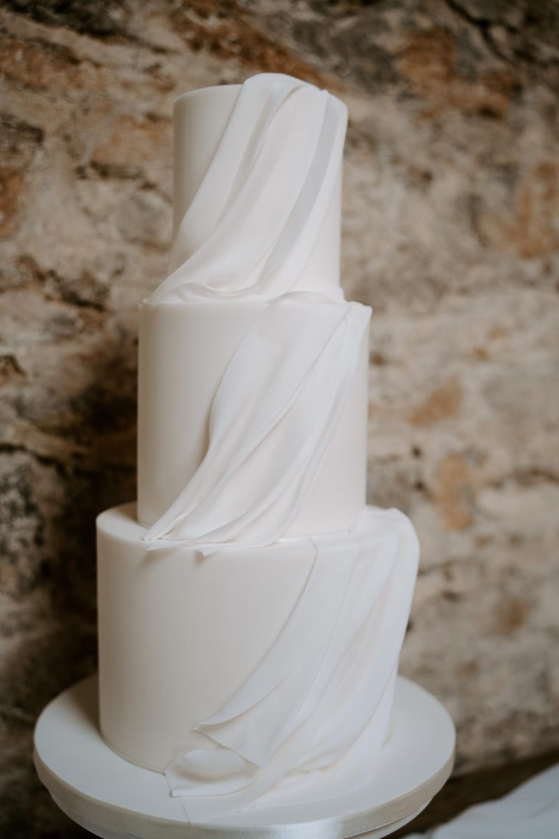 A three-tiered white wedding cake with minimalist draped white fondant decorations stands against a stone wall background.