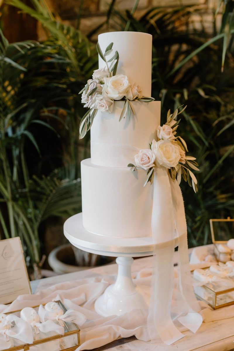 Handcrafted wedding cakes. A three-tier white wedding cake adorned with white roses and greenery, displayed on a white cake stand with a draped white cloth on the table.