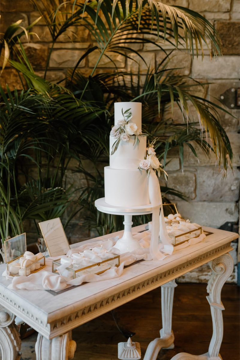 A luxury fondant wedding cake with three-tiers with floral decorations on a white stand, placed on a decorated table with individual dessert boxes, surrounded by greenery.