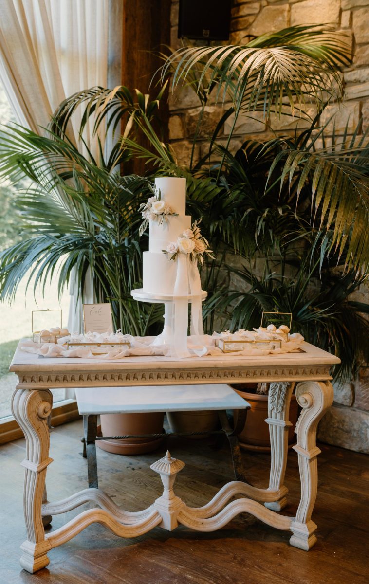 A three-tiered white cake with floral decorations sits on a table surrounded by greenery and a stone wall backdrop.