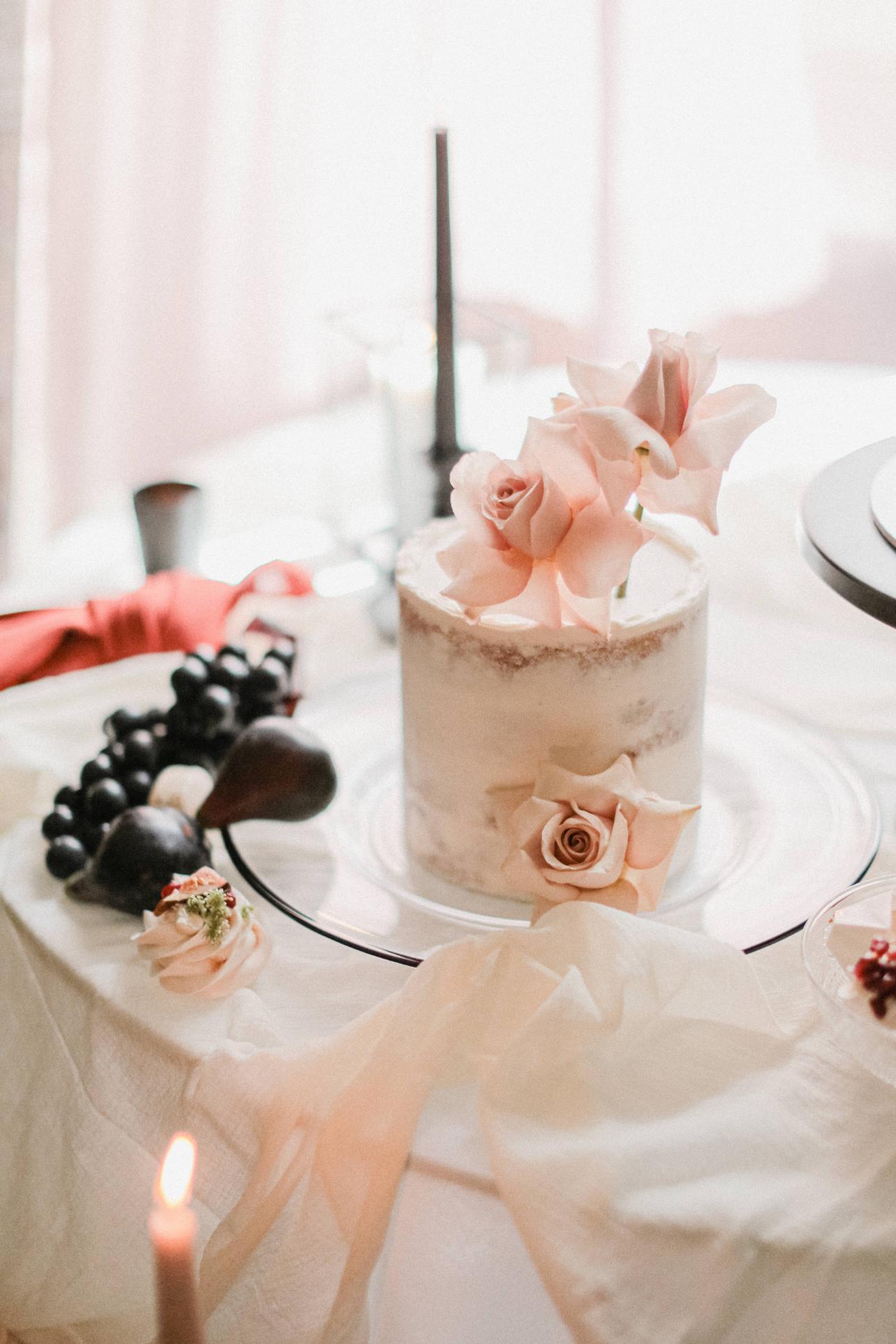 Handcrafted wedding cakes. A small, semi-naked cake adorned with light pink roses sits on a glass plate. The table features a candle, grapes, and decorative elements in the background. Handcrafted wedding cakes