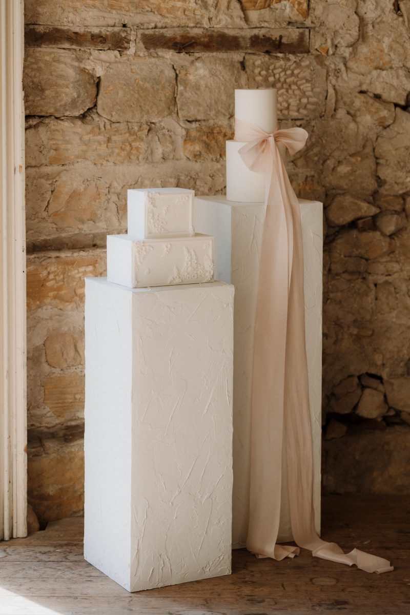 Three white, textured rectangular structures of varying heights are placed against a rustic stone wall. The tallest structure is adorned with a nude-colored ribbon.