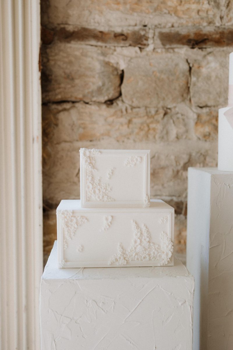 About as elegant as it gets, this white, rectangular tiered wedding cake adorned with delicate floral icing details is beautifully displayed against a rustic stone wall.