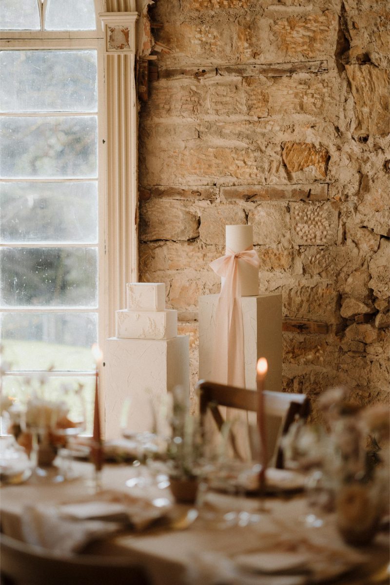 A rustic interior with a brick wall, a large window, and a wooden table setup for dining. On the windowsill, two stacked luxury fondant wedding cakes are decorated, one with a pink ribbon.