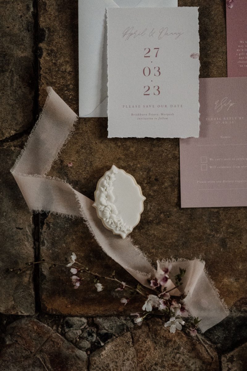 A "Save the Date" card with the date 27.03.23, a decorative item, ribbon, and pink flowers are arranged on a textured brown surface.