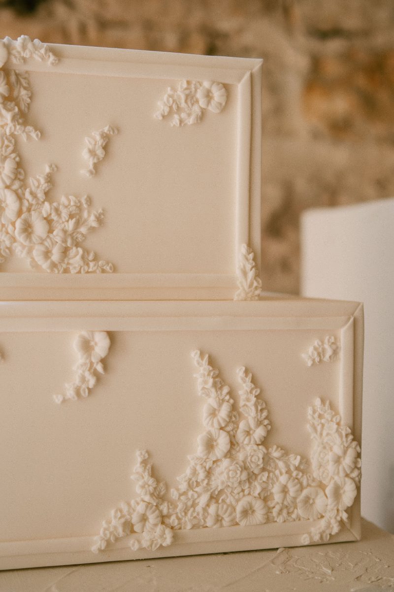 Close-up of a two-tiered white cake adorned with intricate floral decorations against a blurred stone wall background.