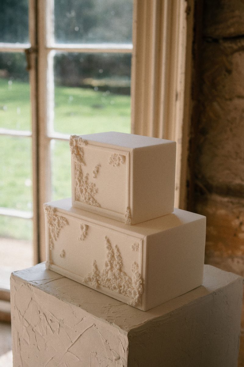 A two-tiered rectangular cake with minimalist design and subtle textured decorations is placed on a pedestal near a window with a scenic outdoor view.
