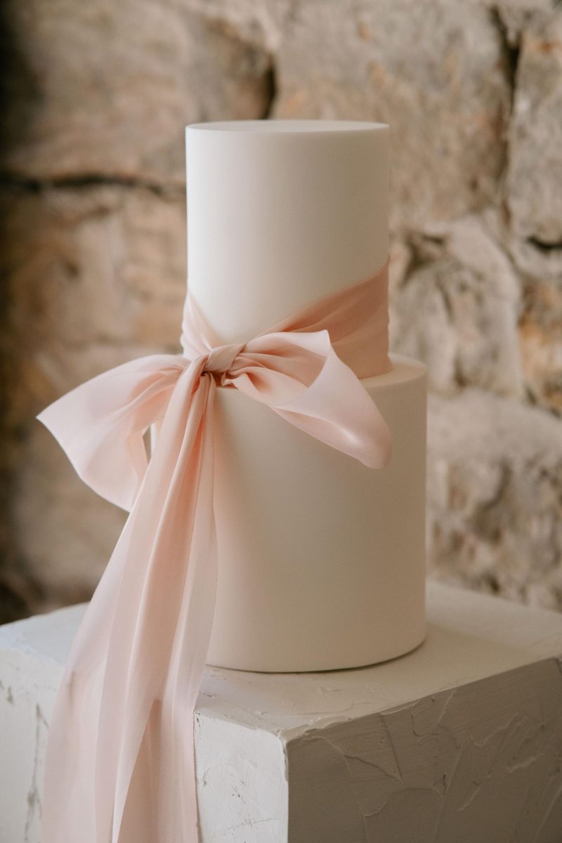 Wedding cake in South Shields. Minimalist two-tier white wedding cake adorned with a wide, pale pink ribbon bow, set against a stone wall backdrop.