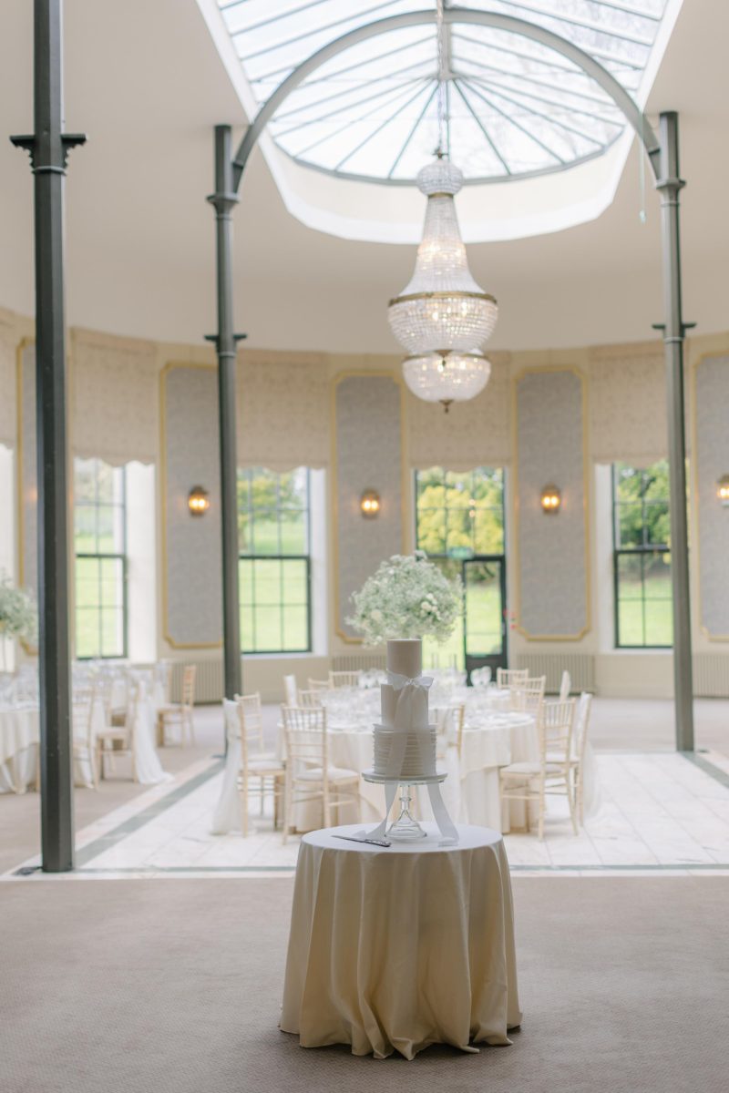 A wedding cake on a round table is in the foreground, with an elegantly decorated reception area featuring chandeliers, round tables, and large windows in the background.