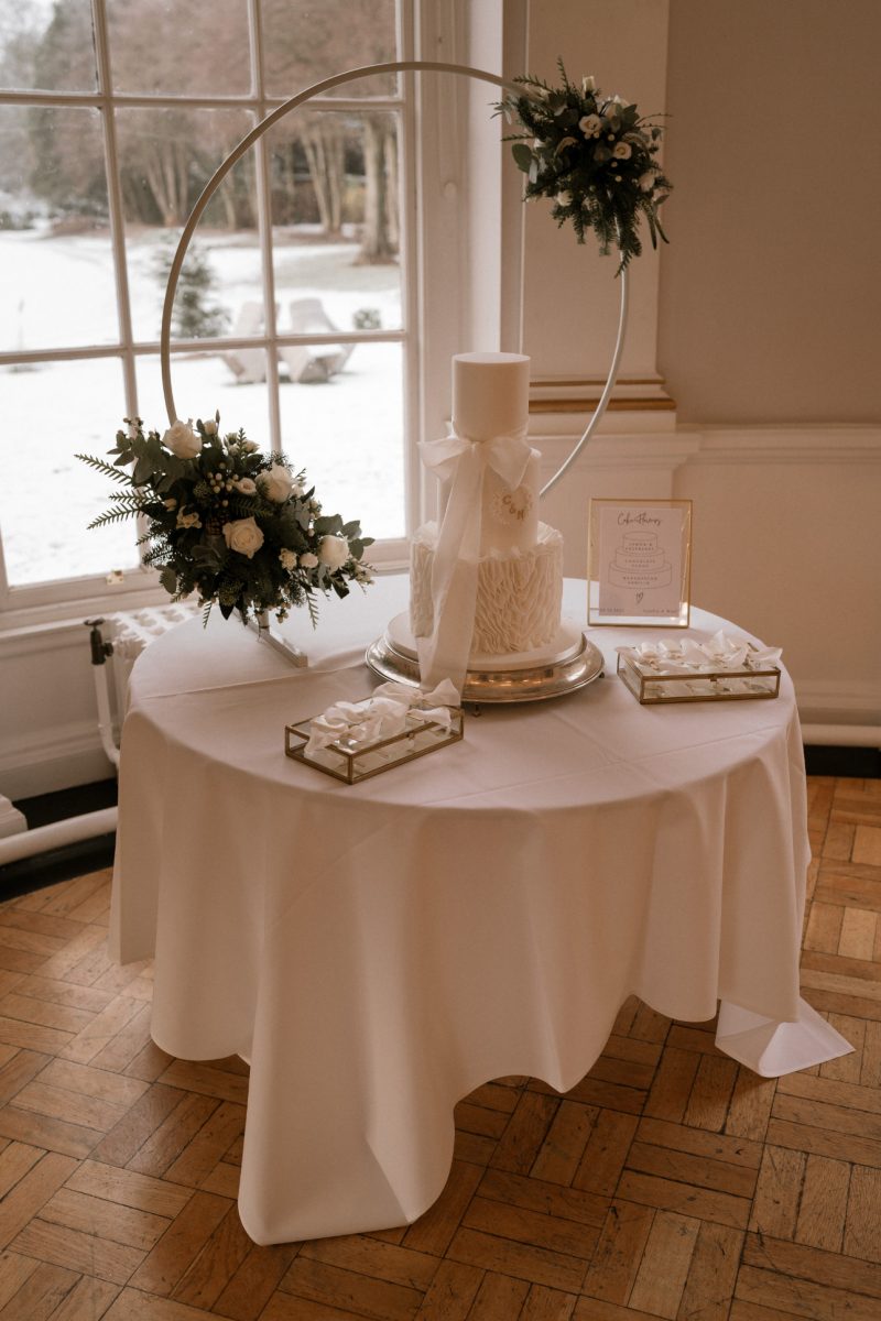 A white frosted cake is displayed on a round table covered with a white cloth. The cake is accented with greenery. The window behind reveals a snowy outdoor scene. Two trays of treats and a card are also on the table.