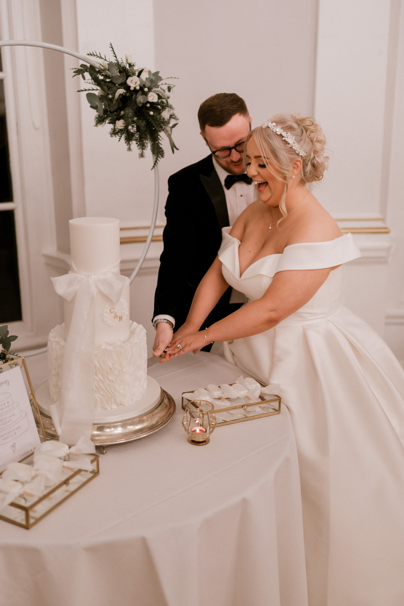 A bride and groom in formal attire cut a white, multi-tiered wedding cake on a decorated table. Created by The Little Cakery, a wedding Cake Designer North East England.