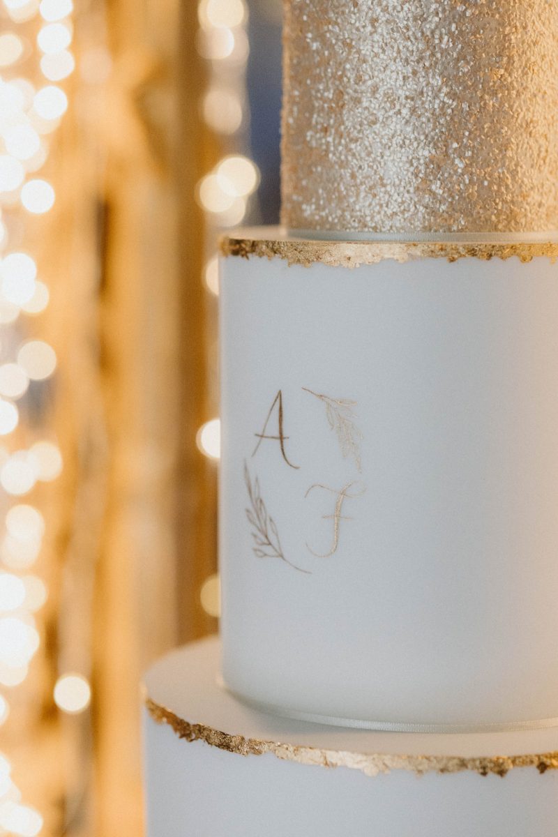 Close-up of a white and gold tiered wedding cake. The middle tier has an "A" and "F" monogram with ornate designs, and the top tier is covered in shimmering gold. Warm, glowing lights are blurred in the background.