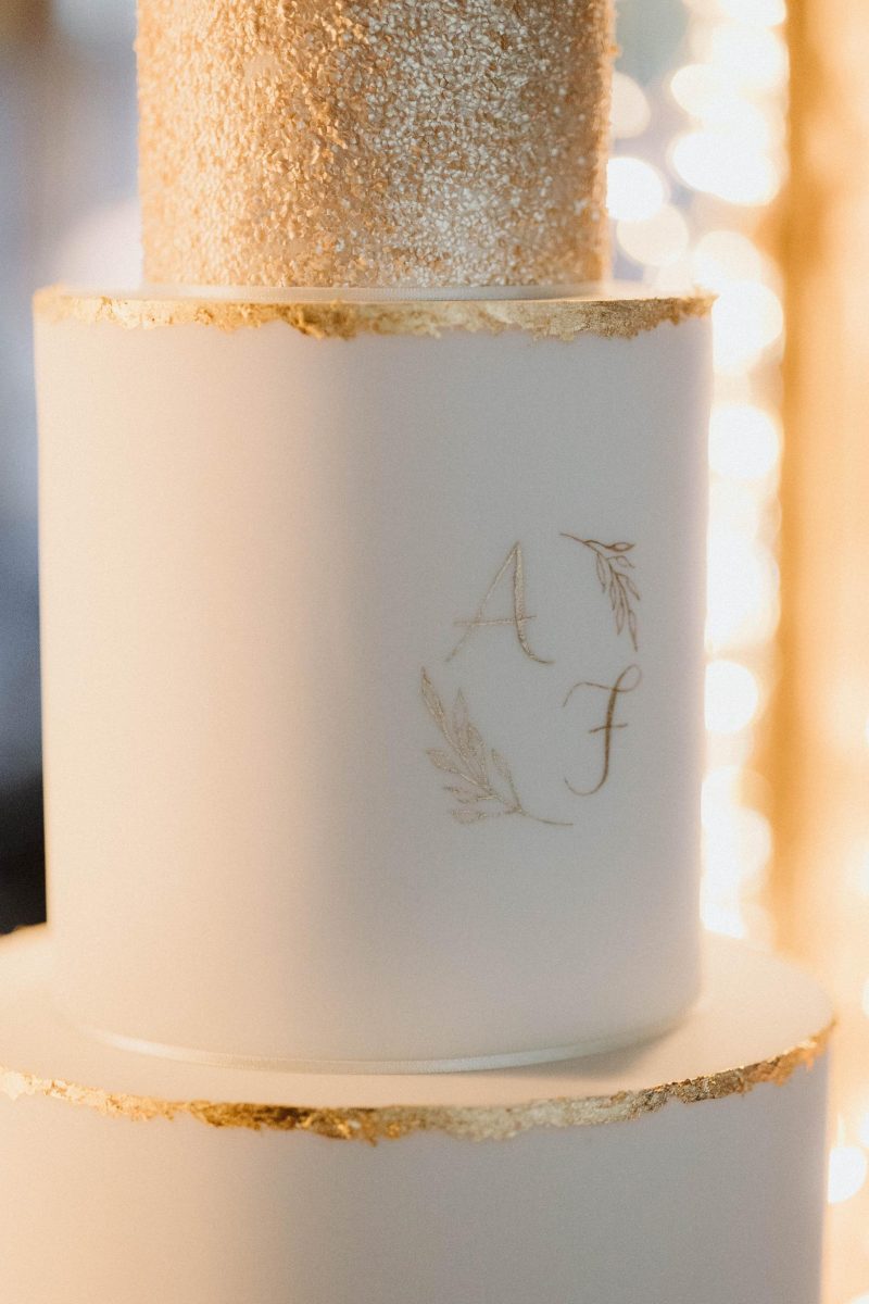 Close-up image of a multi-tiered white cake with gold leaf accents and the initials "A" and "F" inscribed on one tier, surrounded by a decorative laurel wreath design.