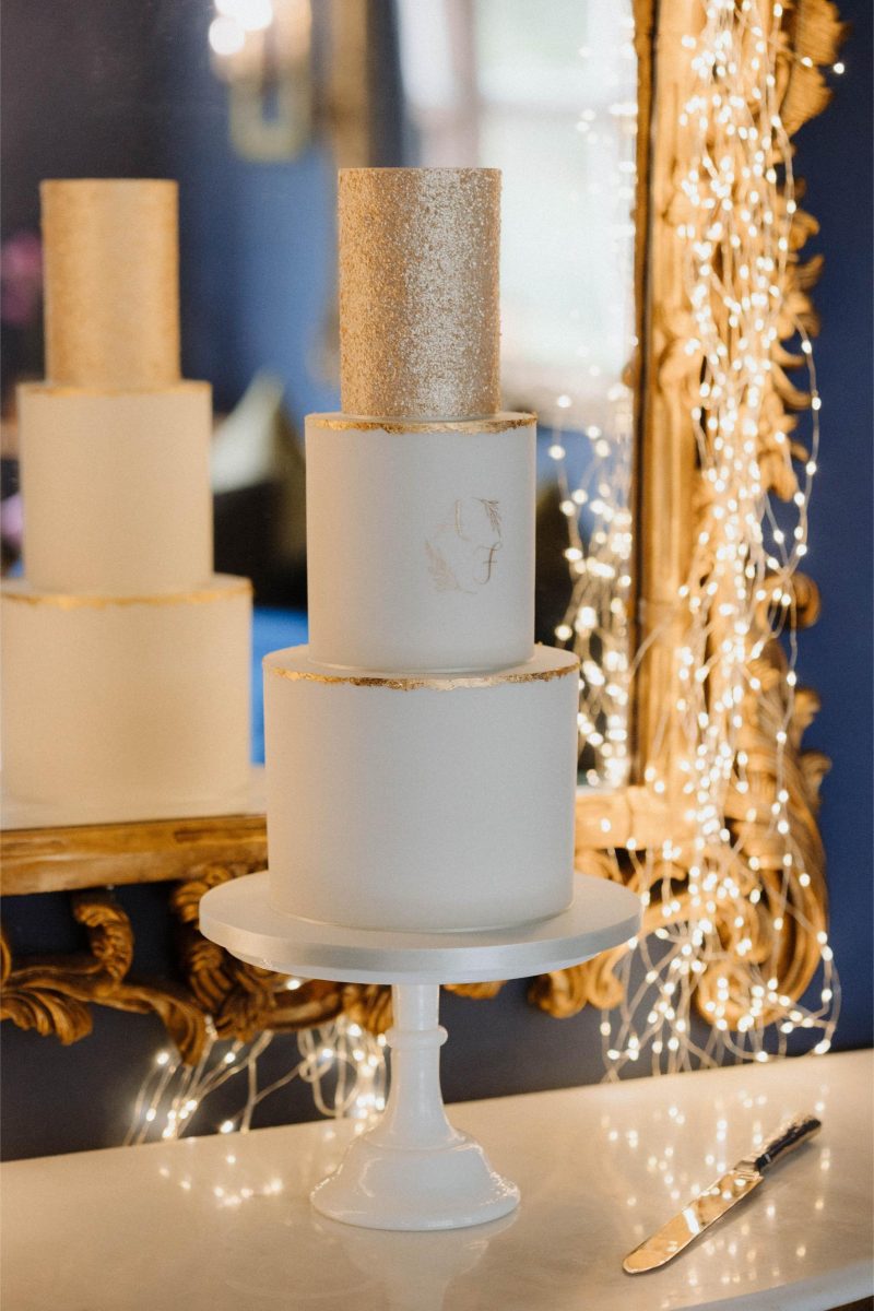 A three-tier white cake with gold accents and a glittery top tier, displayed on a white pedestal, with decorative lights and a gold frame mirror in the background. A cake knife rests nearby.