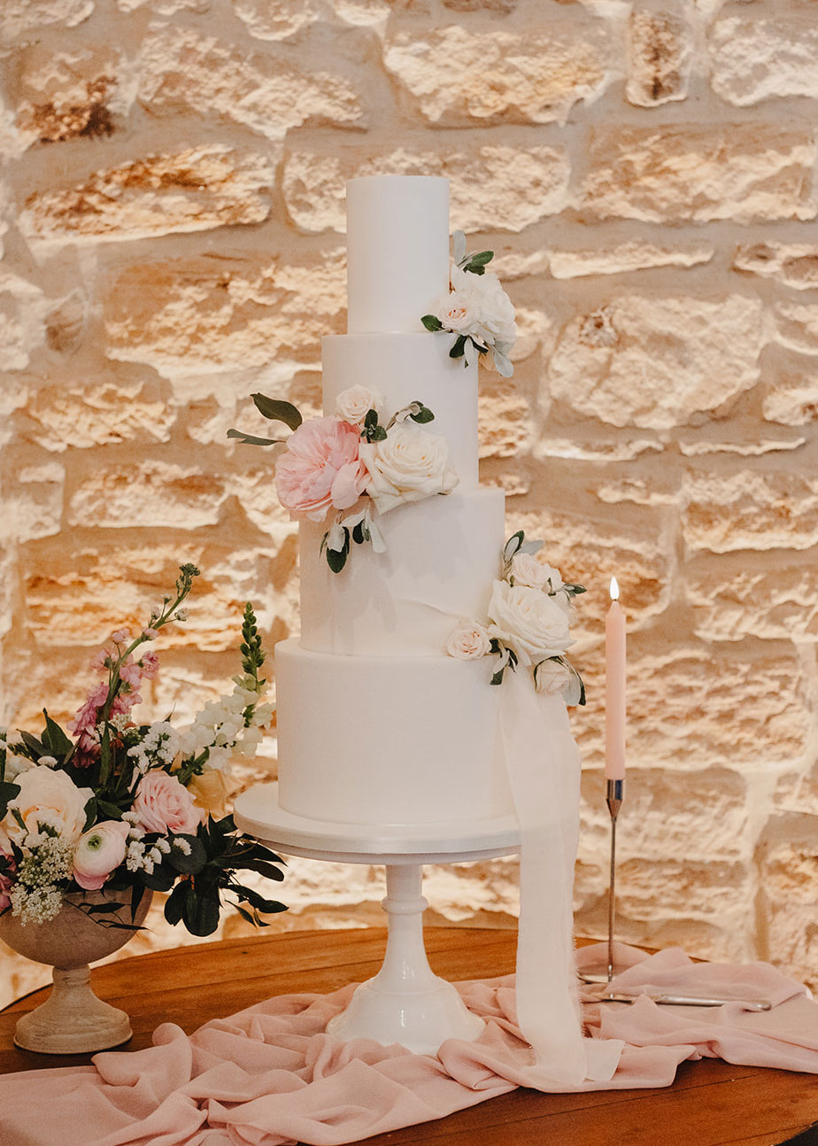 A luxury fondant wedding cake with four-tiers in white, resembling delicate artwork, is decorated with pink and white flowers and greenery. Displayed on a white stand, this edible masterpiece is accompanied by a candle and flower arrangement on a pink cloth, creating an exhibition of elegance.