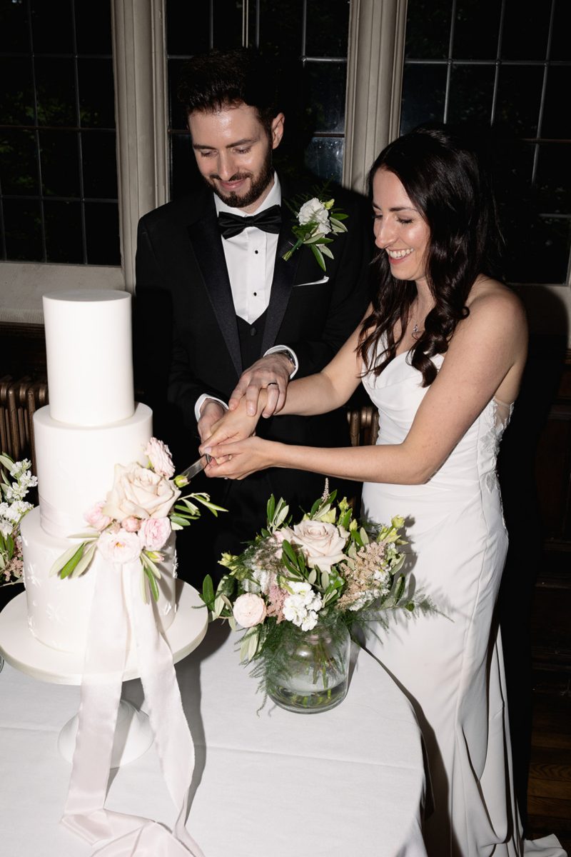 A couple in formal attire is cutting a tiered white cake decorated with flowers at a wedding reception. They are smiling and standing close to each other.