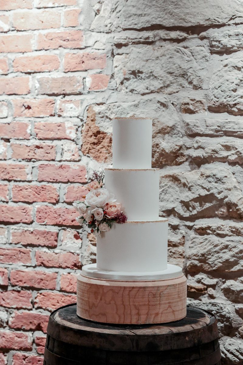 A three-tier white wedding cake with a floral arrangement is displayed on a wooden pedestal against a rustic brick and stone wall.