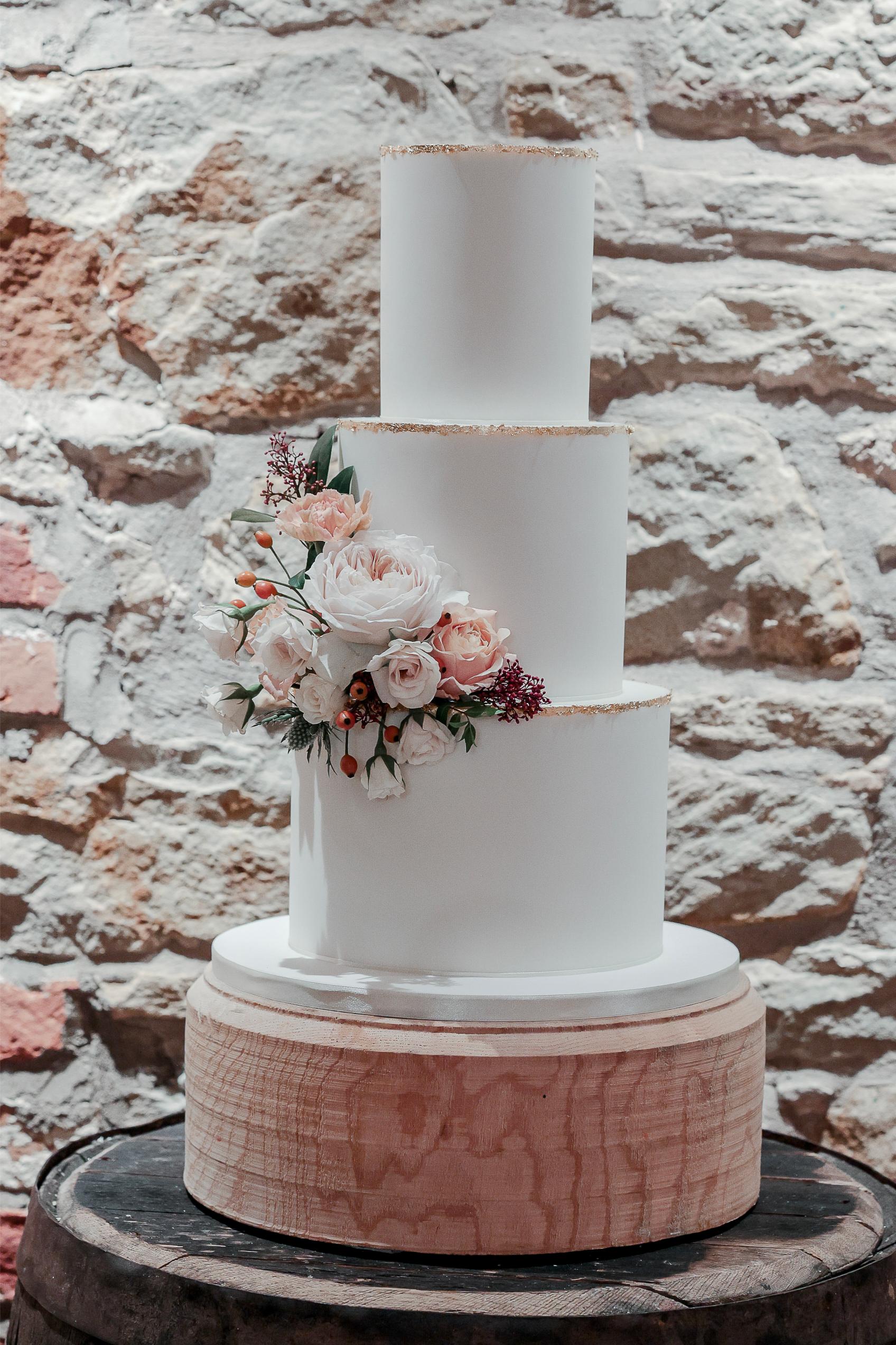Handcrafted wedding cakes. Three-tier white wedding cake with floral decorations, displayed on a wooden stand against a rustic brick wall.