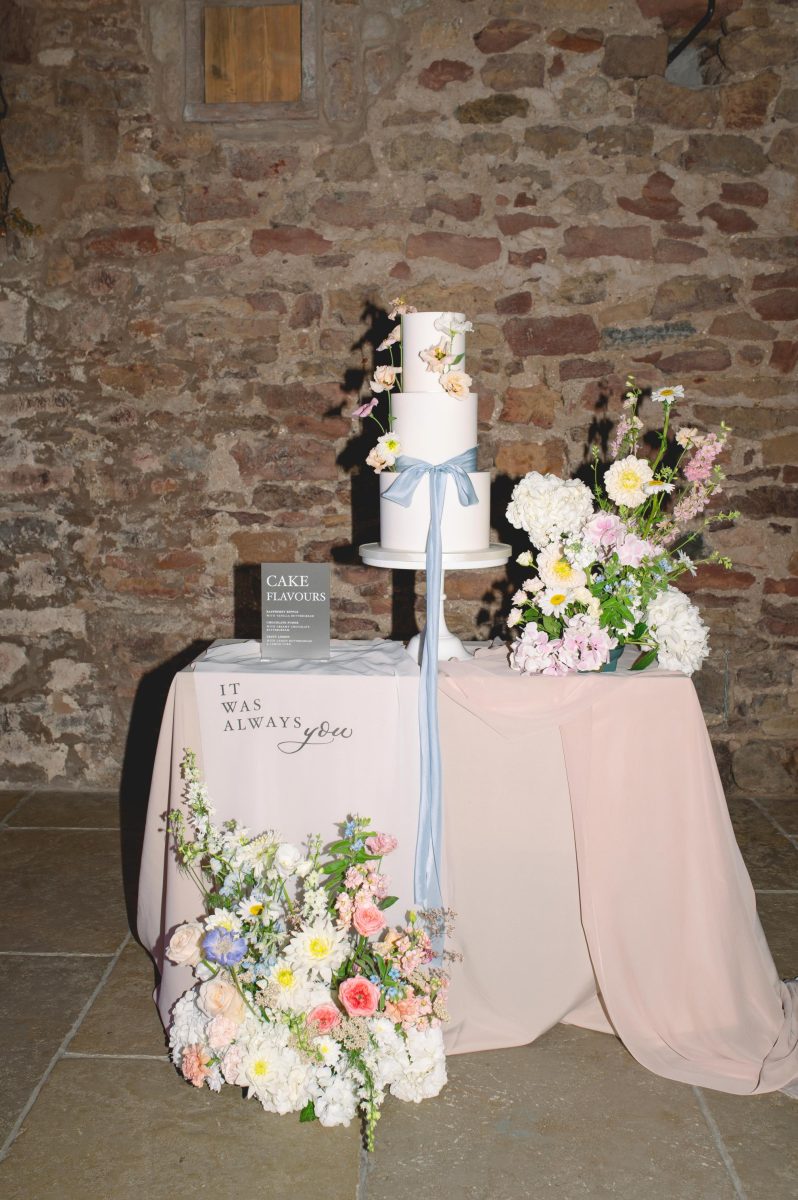 A two-tier white wedding cake with floral decorations and blue ribbon on a table with light pink cloth, surrounded by floral arrangements. A sign reads "It was always you." A book titled "Cake Flavours.