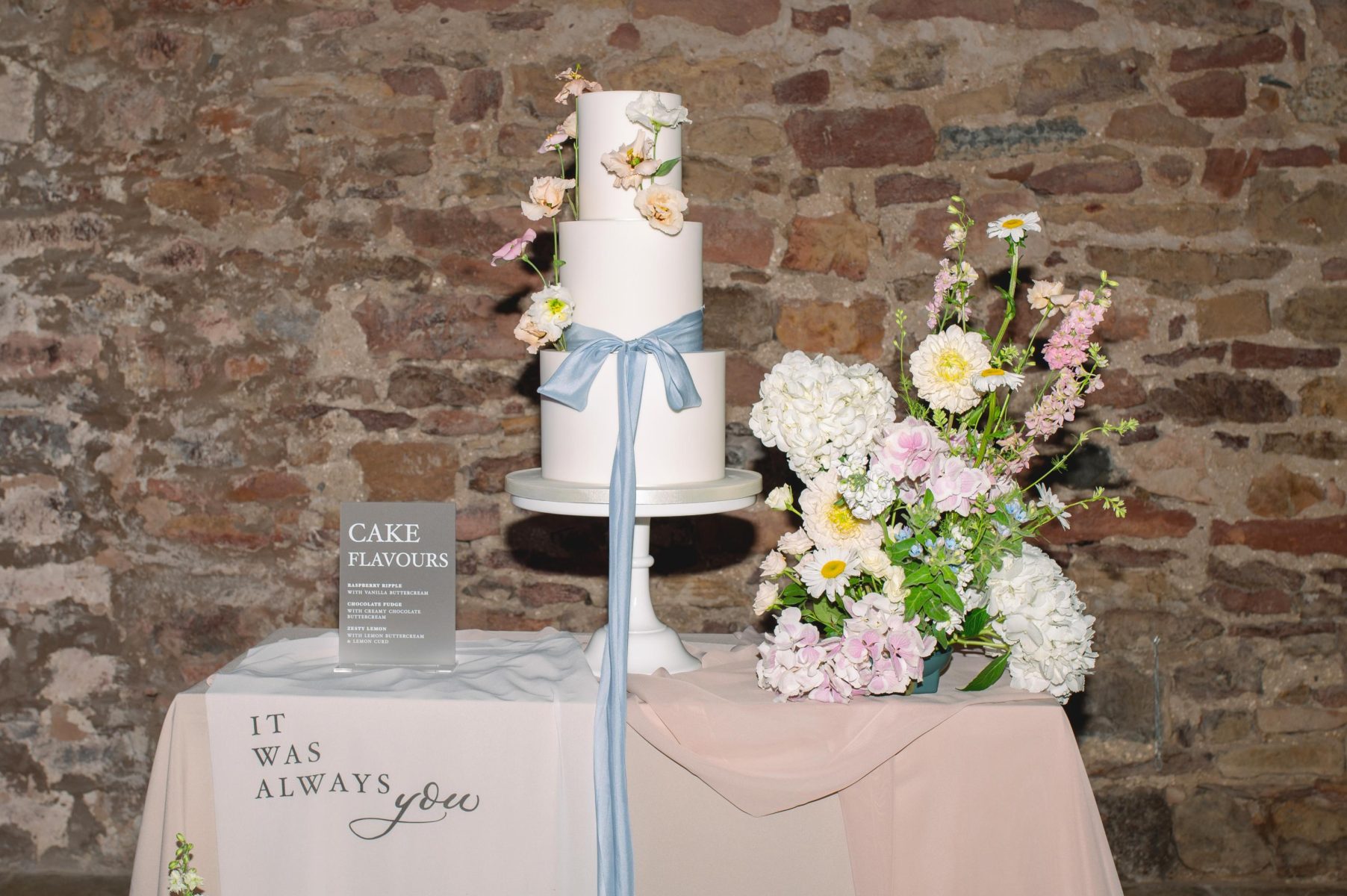 A three-tiered white wedding cake adorned with flowers and a blue ribbon stands on a table. Nearby, a sign lists cake flavors. A floral arrangement and a cloth with "It was always you" are also displayed.
