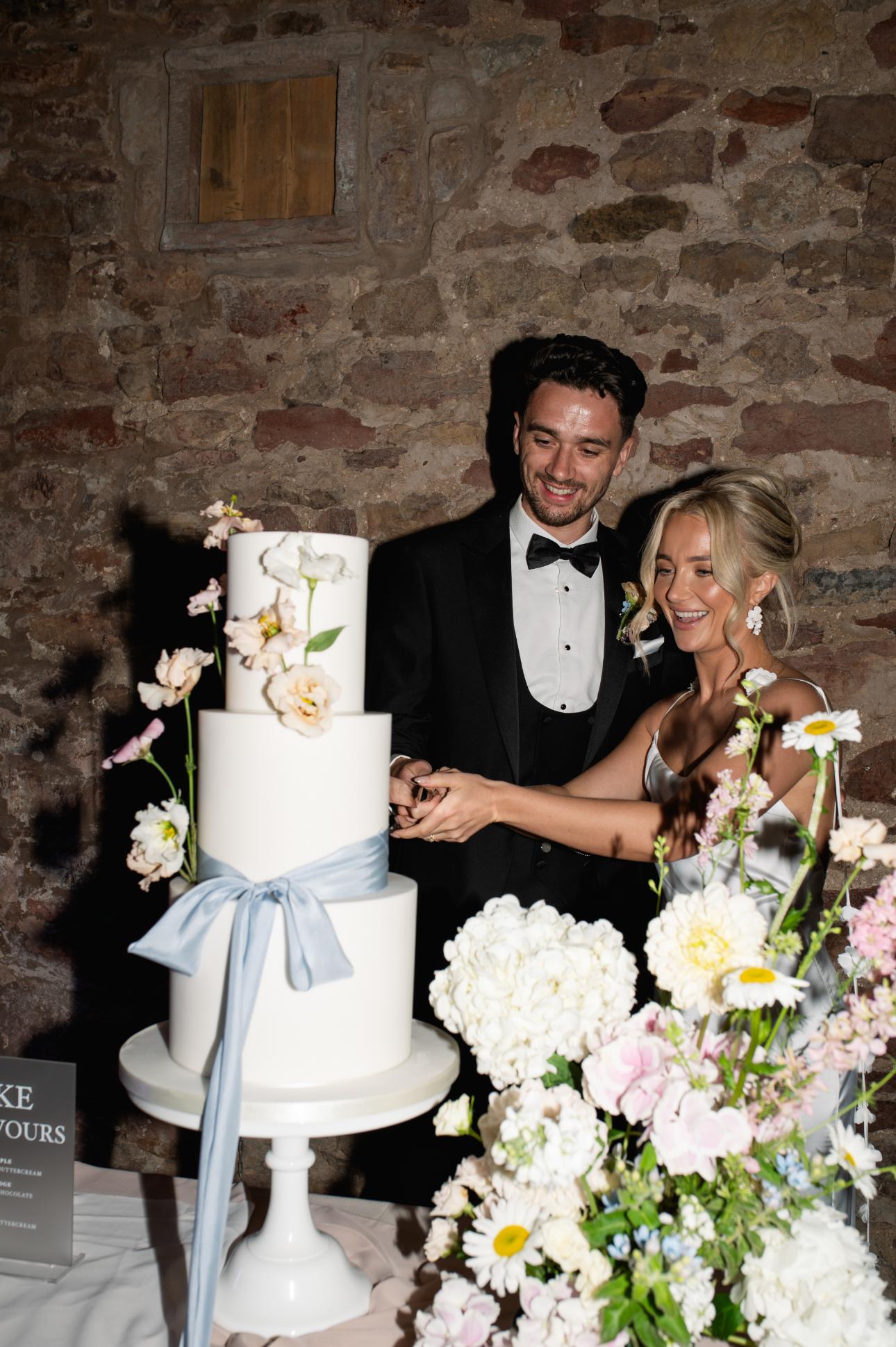 A couple in formal attire cut their luxury fondant wedding cake decorated with flowers and a blue ribbon in front of a brick wall.