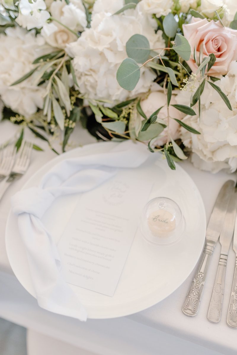 A white plate with a folded napkin, a small clear container labeled "Bride" holding a small item, and a printed menu sit on a table adorned with white and pink flowers.
