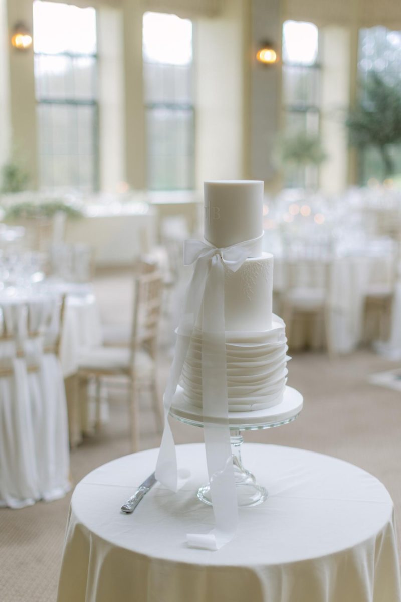 A three-tier white wedding cake with a white ribbon on a glass stand, placed on a round table with a white tablecloth in a banquet hall with white chairs and tables in the background.