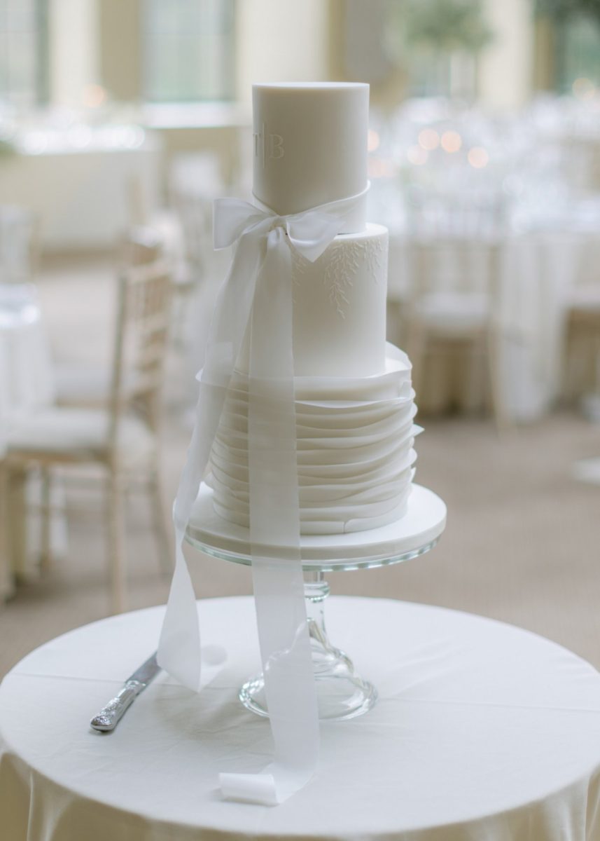 A white, three-tiered wedding cake with a large ribbon bow on top stands on a round table, with a knife placed beside it. The background features blurred chairs and tables in a light-filled room.
