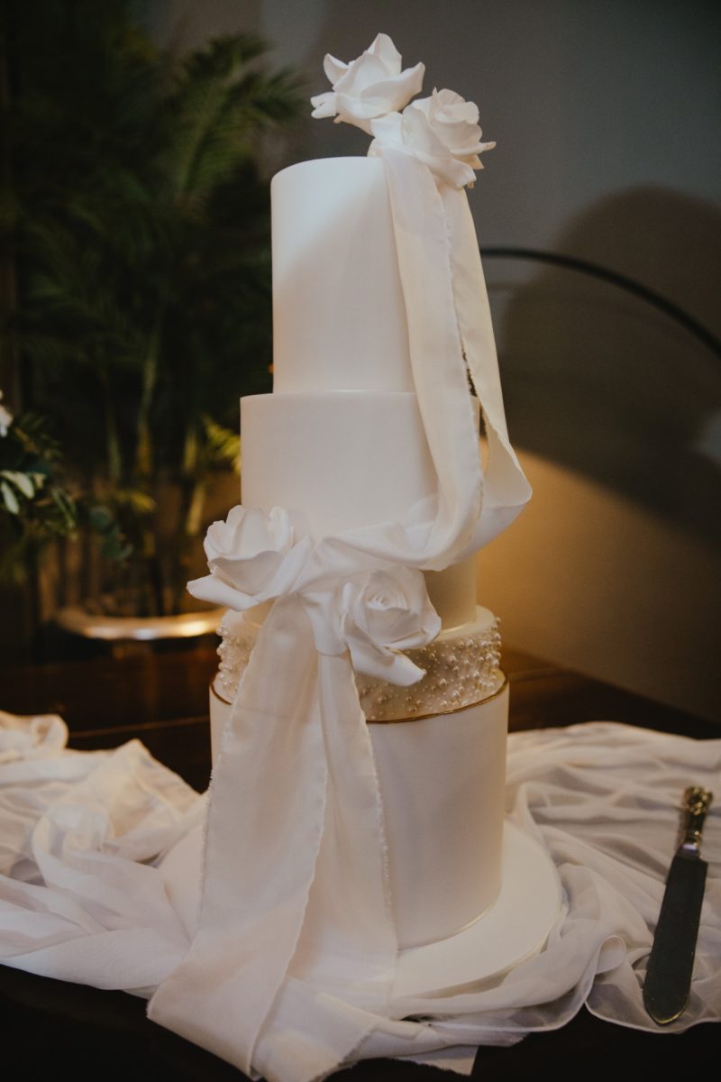 A three-tier white wedding cake decorated with large white fondant flowers and draped with white ribbon, set on a table with white fabric.
