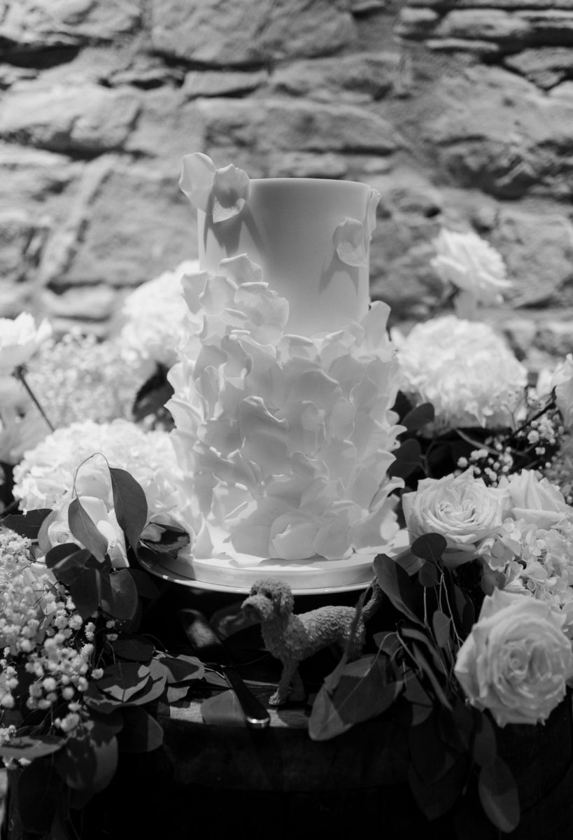 A two-tier white cake decorated with delicate floral elements stands like a gallery centerpiece amid an arrangement of flowers and foliage, with a small dog figurine placed at its base.