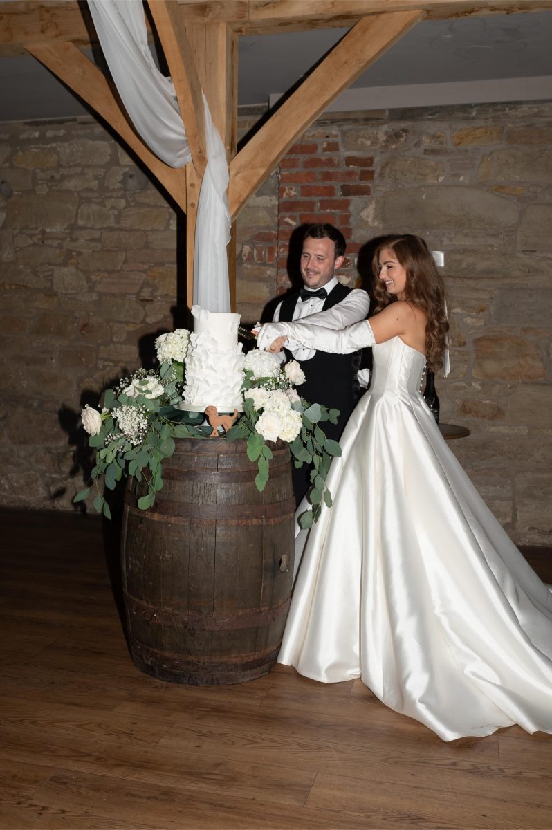 A bride and groom in wedding attire cut a cake on a barrel, decorated with flowers, in a rustic venue with stone walls and exposed wooden beams.