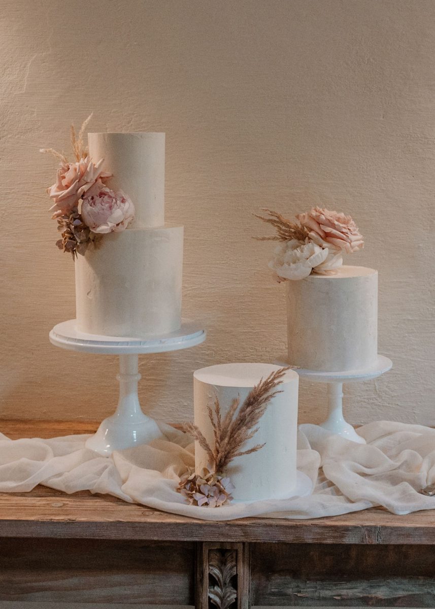 Three minimalist white cakes on stands, each adorned with sparse floral decorations, positioned on a wooden table draped with white fabric.