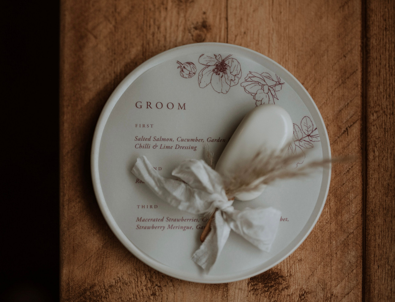 A wedding menu labeled "Groom" featuring three meal courses, decorated with floral illustrations and a white oval piece wrapped in a ribbon, placed on a wooden table.