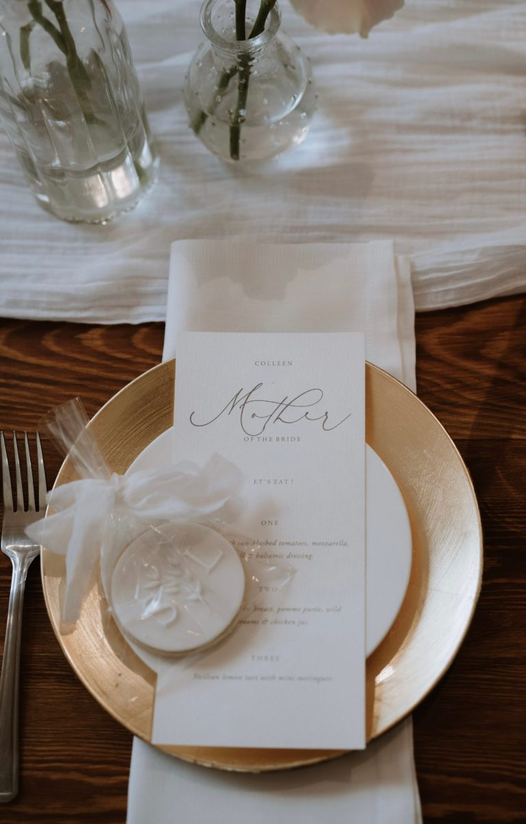 A wedding menu card placed on a white napkin atop a gold charger plate, with a clear-wrapped cookie favor and silverware beside it.