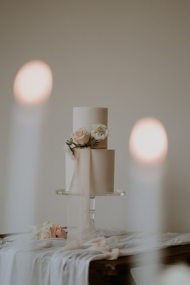 Wedding cake in South Shields. A two-tiered white cake adorned with soft pink roses and white flowers, placed on a glass stand, with two out-of-focus candles in the foreground.