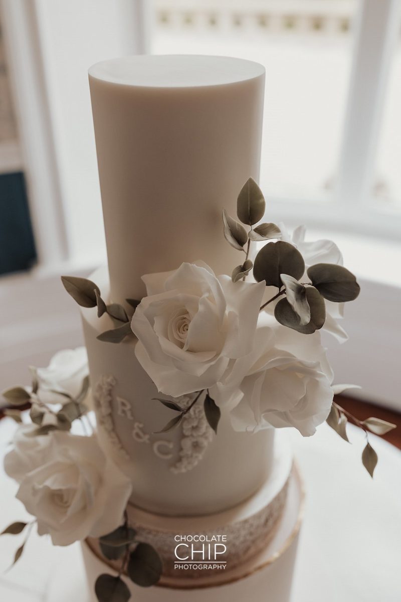 A three-tier white wedding cake adorned with white roses and green foliage stands like an artwork in a gallery. The middle tier showcases the initials "R & C." This beautiful creation is placed on a table near a window, basking in natural light.