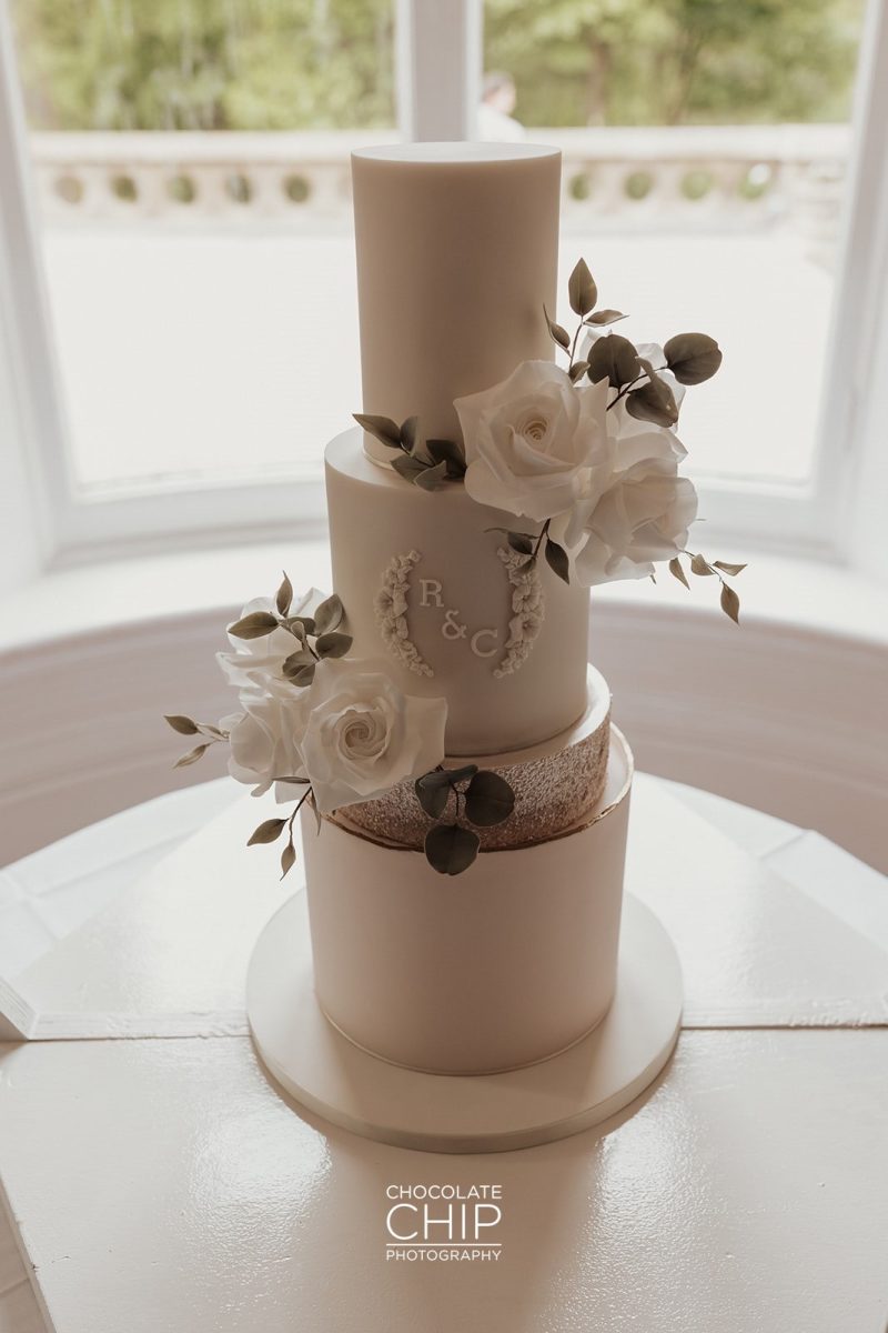 Three-tiered white wedding cake decorated with white roses and green leaves, featuring the initials "R & C", showcased like an art piece in a gallery. The cake is placed on a round table by a window.