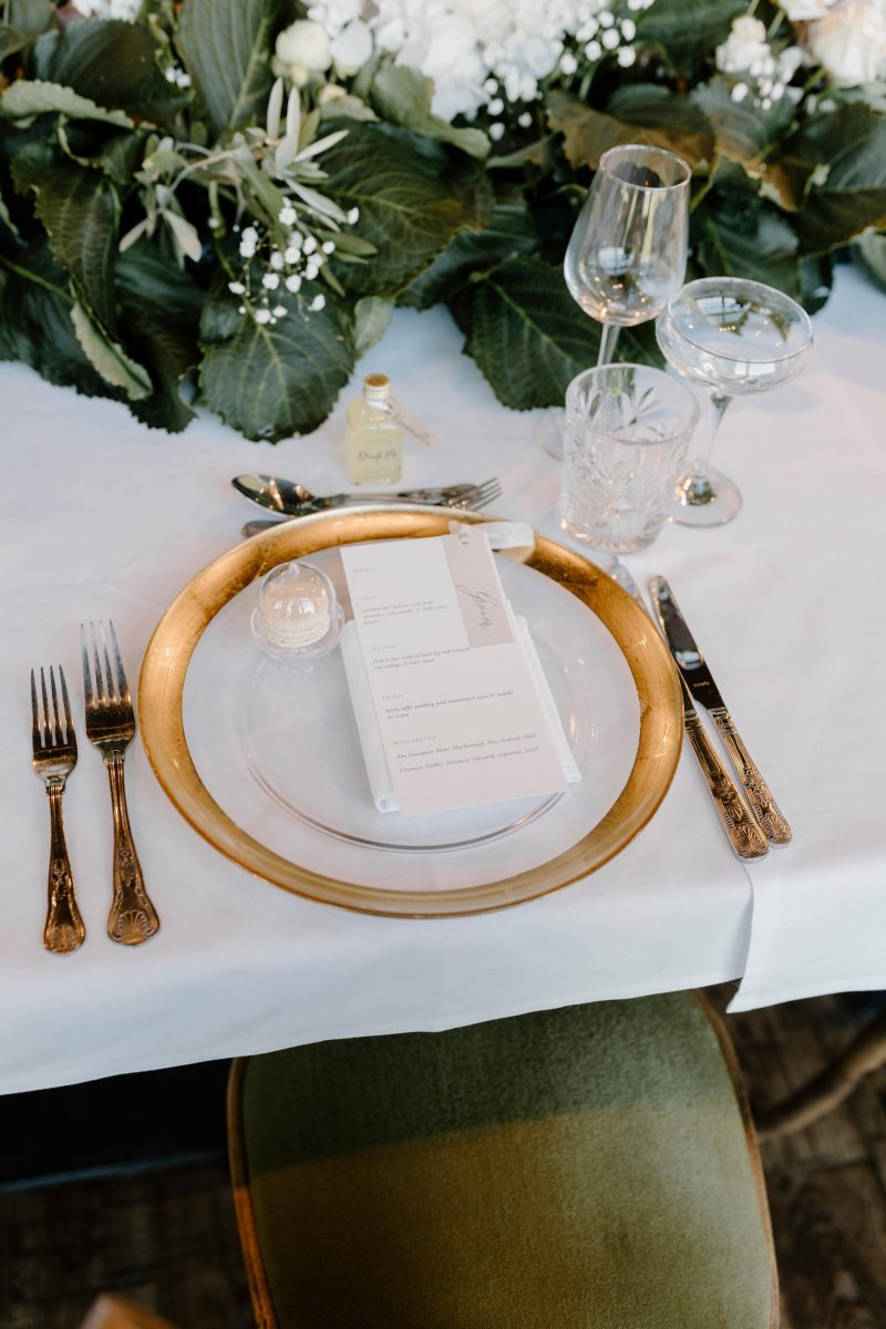 A dining table setting features a glass plate on a gold charger, a menu card, and cutlery on either side. Above the plate are a wine glass, water glass, and a decorated greenery centerpiece.